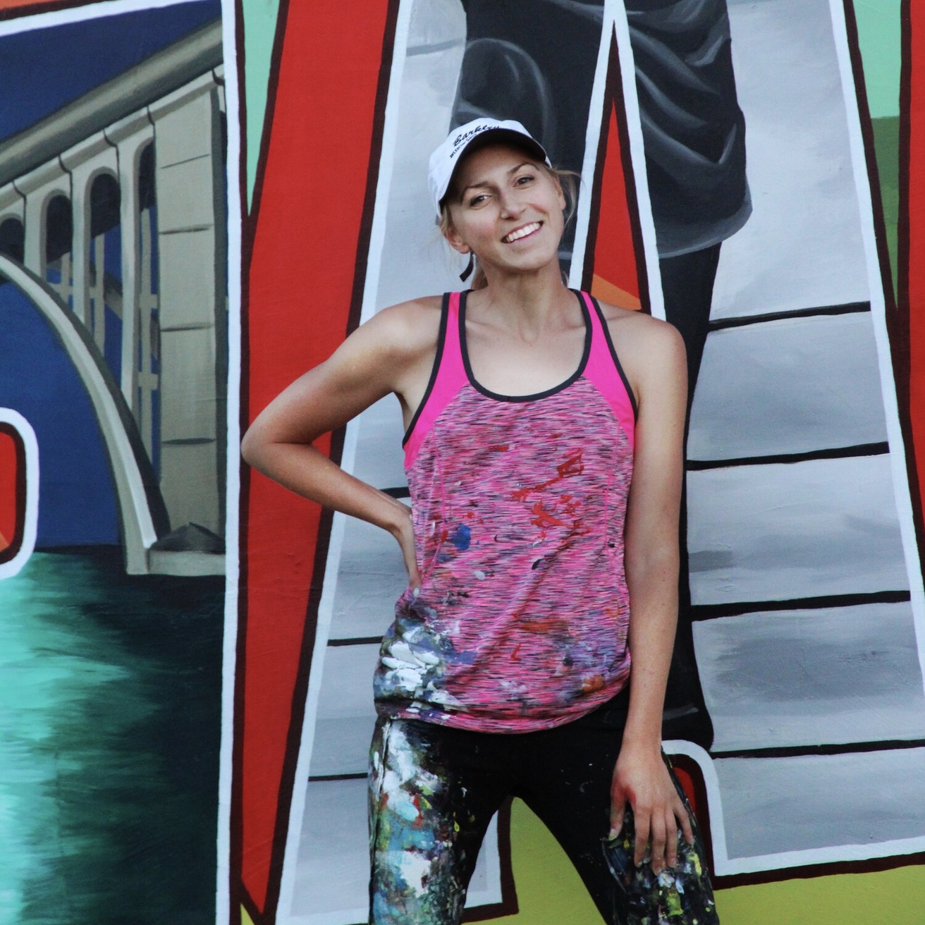 Young woman wearing a hat and splattered with paint standing in front of a mural and smiling.