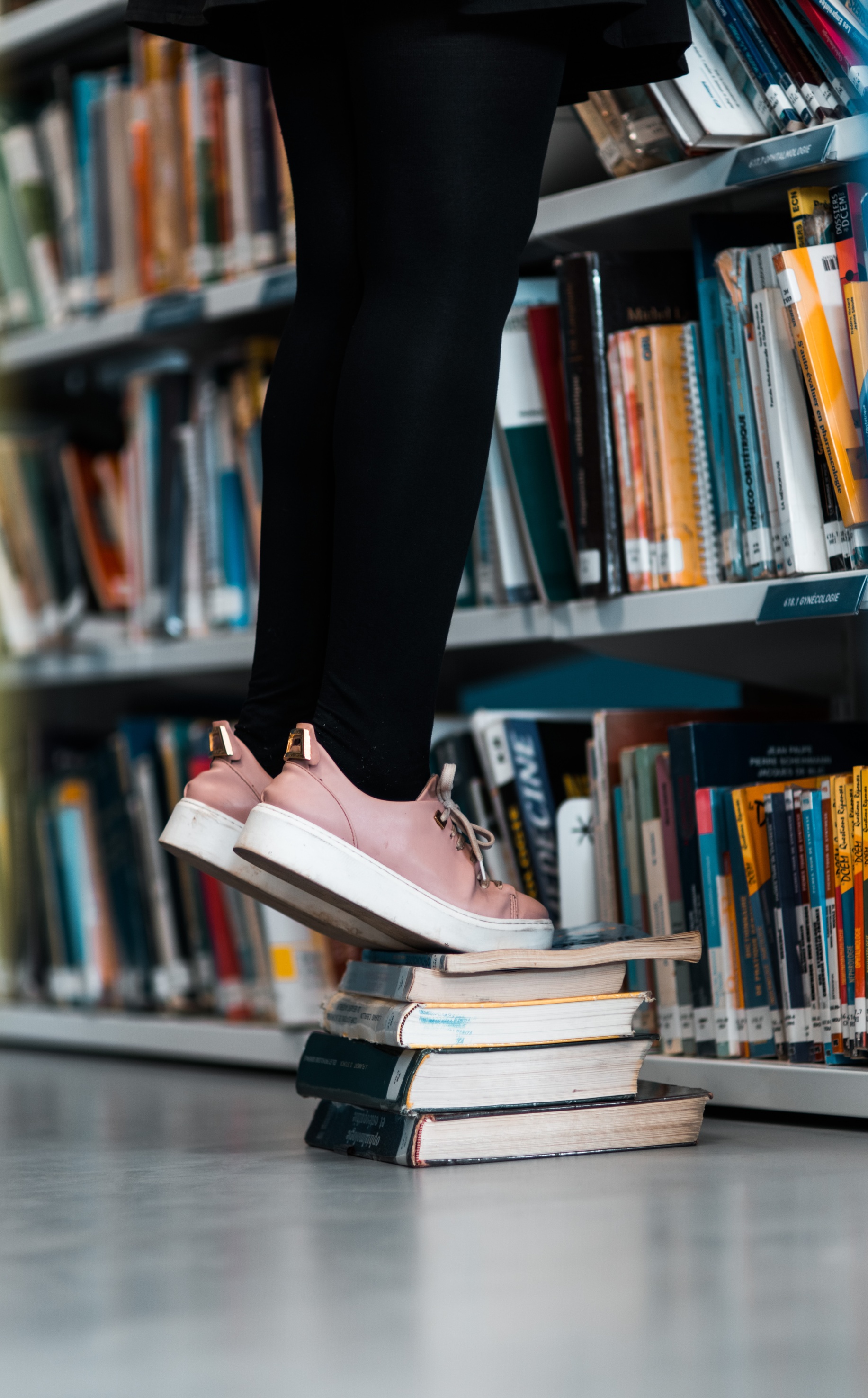 lower legs of person wearing black tights and canvas sneakers standing on a stack of books