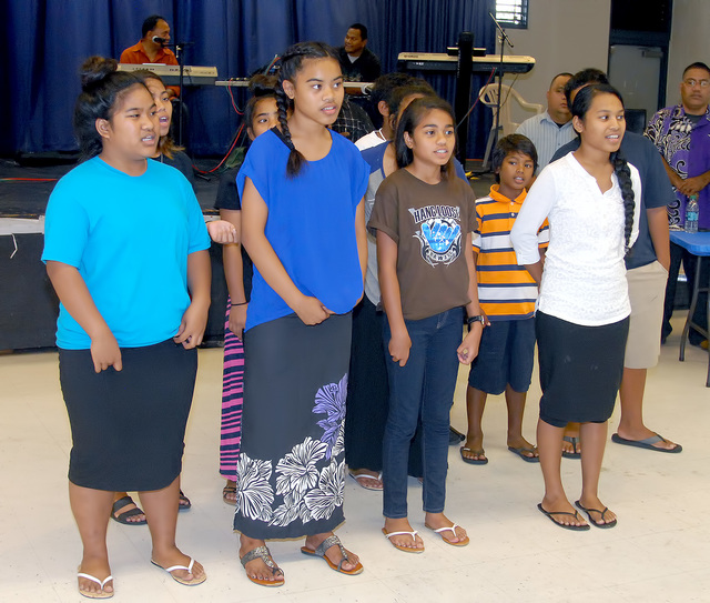 A group of five school-aged people with brown skin stand in a row and look to the side.