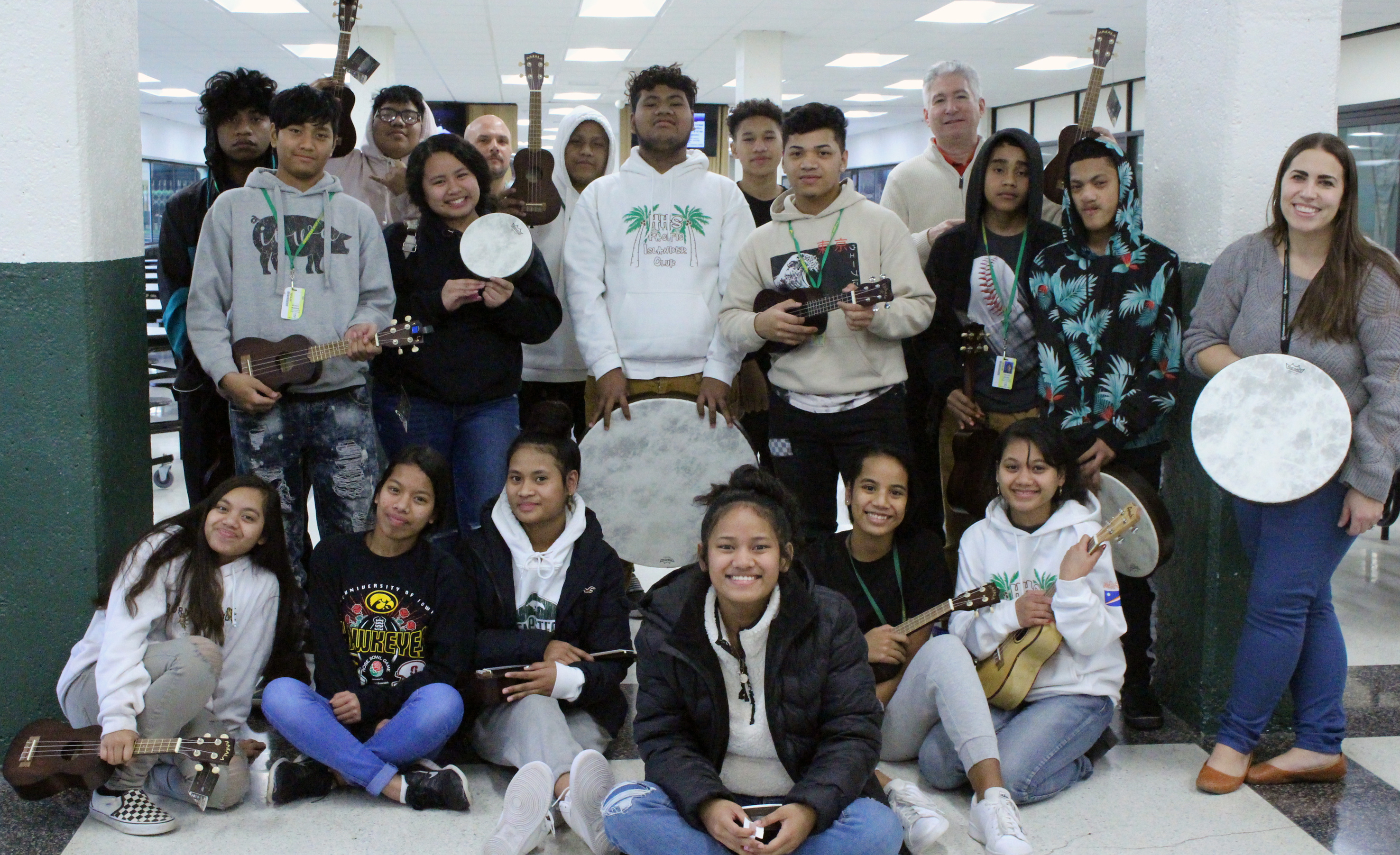 A large group of students pose in the hallway of a school with lockers around them. They are smiling and several are holding musical instruments.