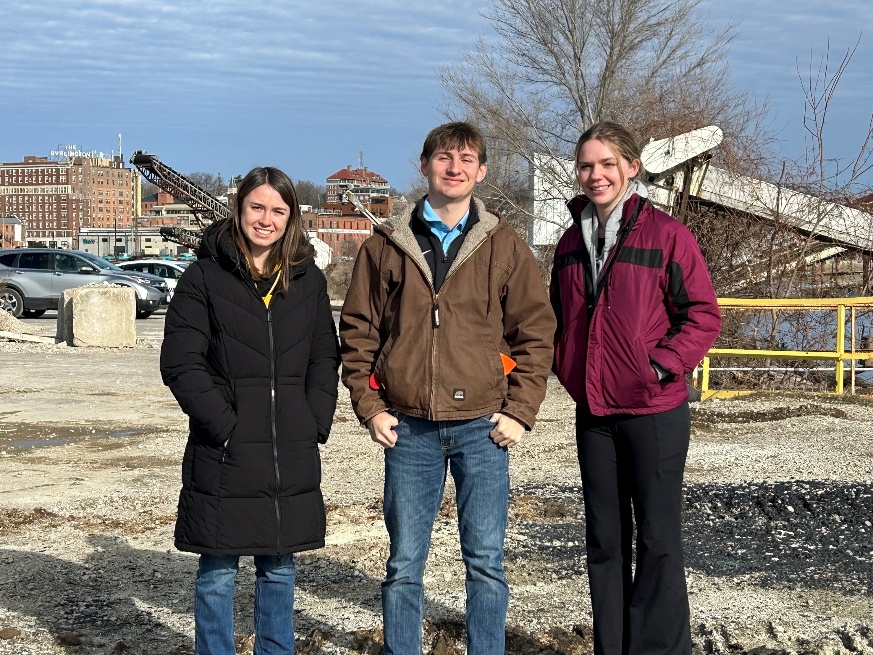 three young people in winter coats standing outside on a sunny day with an industrial site behind them.