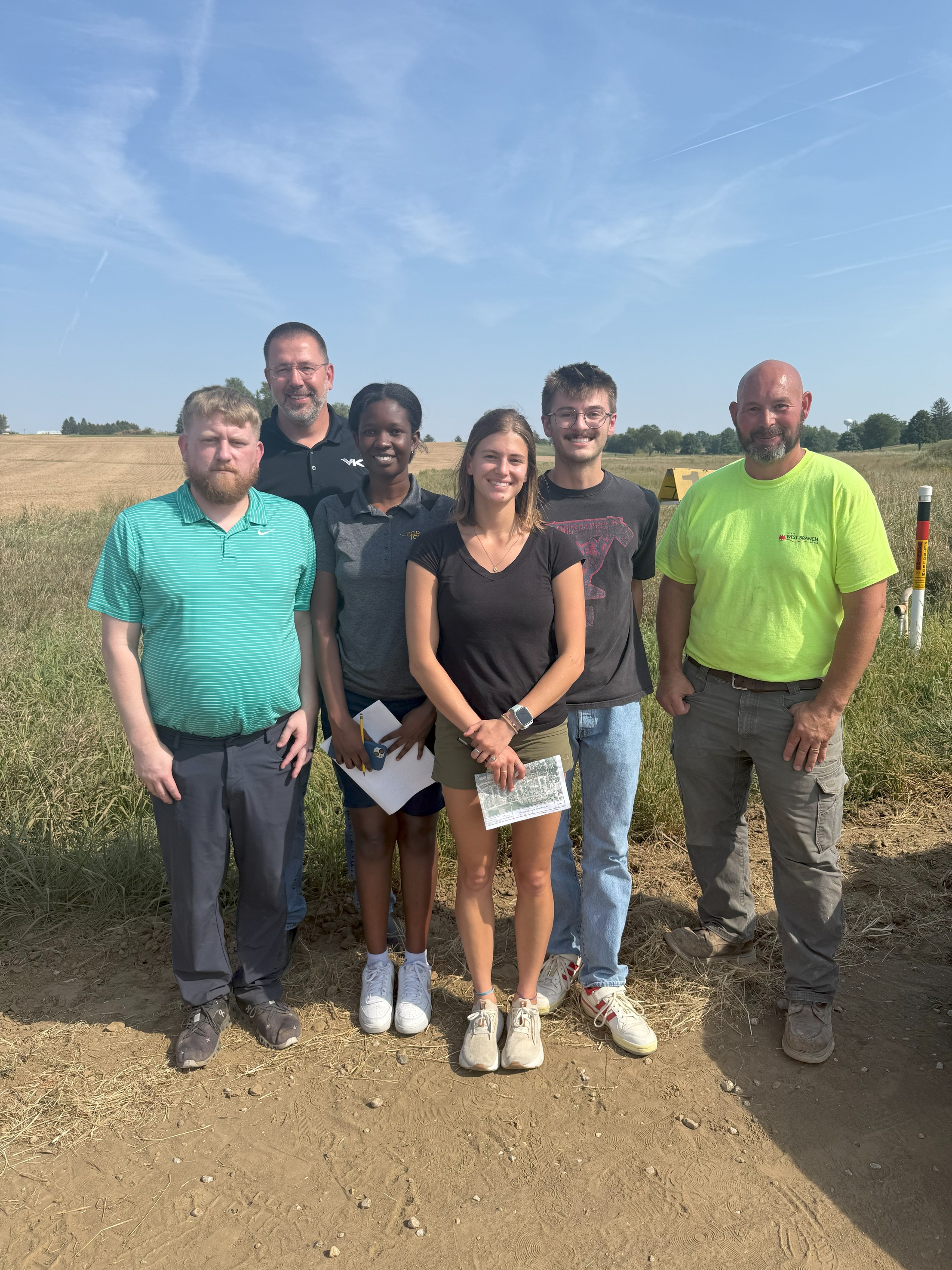 Six people stand on a sunny day in front of a corn field. The three middle people are holding paper maps. The people on each end are wearing West Branch t-shirts.