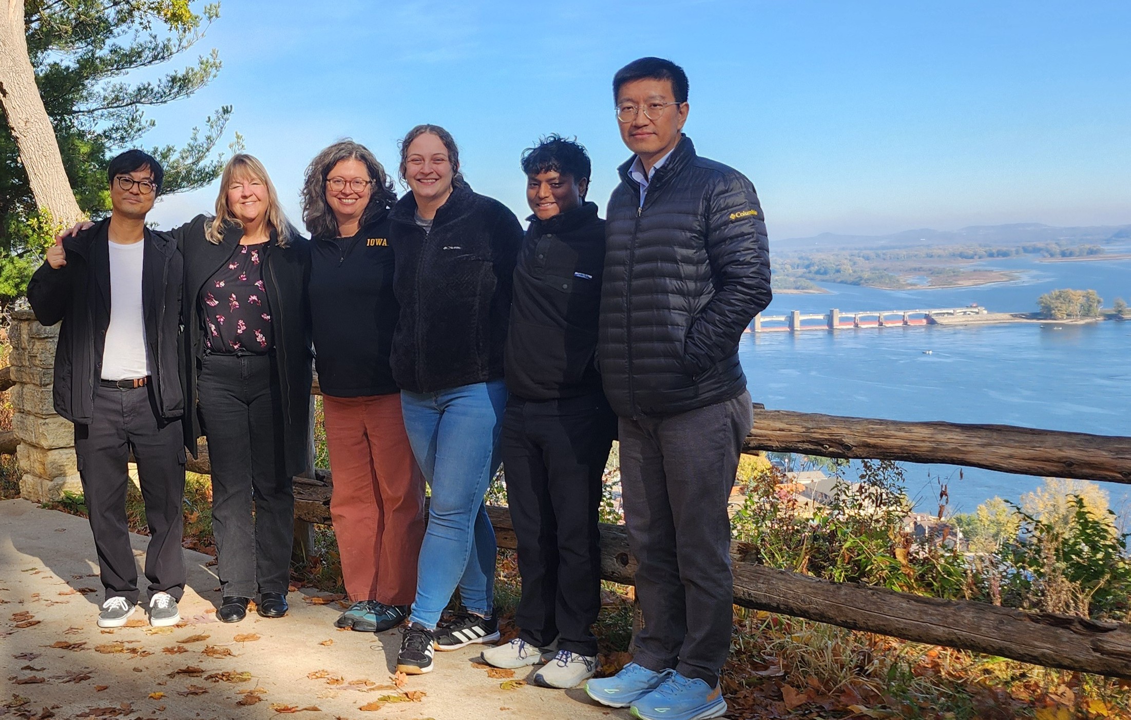 A group of six people stand arm in arm smiling in front of the Mississippi river