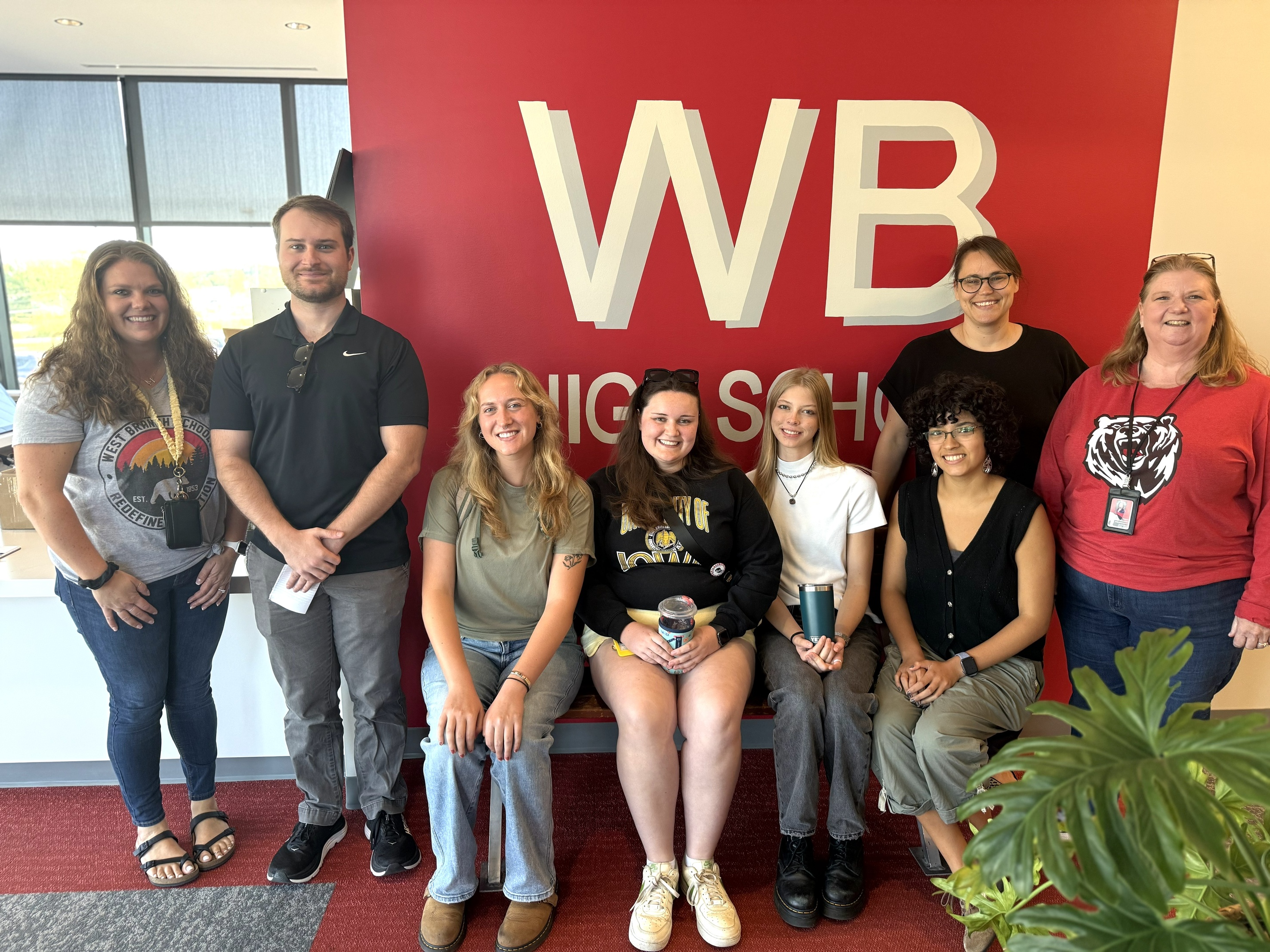 A row of seven people stand and sit smiling in front of a red wall with white letters that says WB