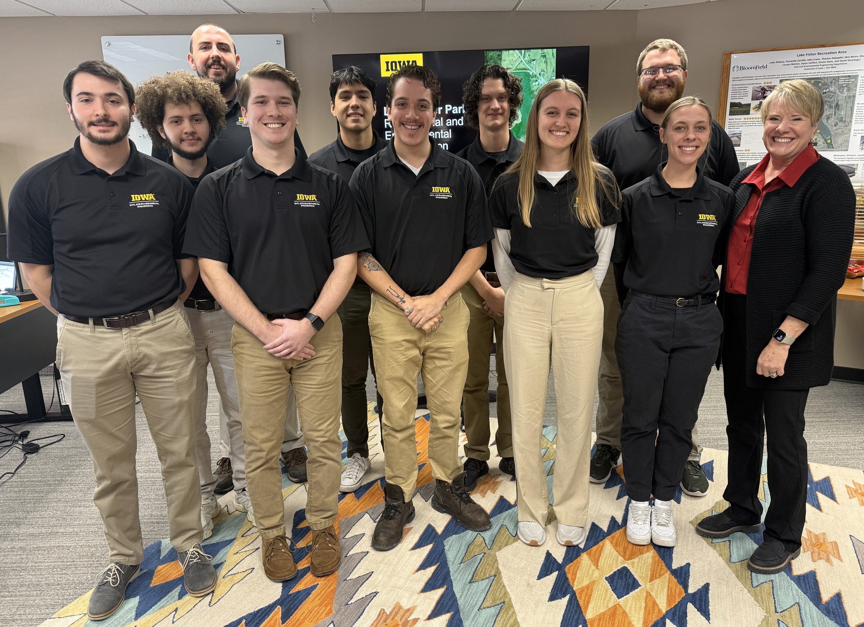 A group of students wearing black polo shirts with the Iowa logo stands with a woman in a red shirt. Everyone is smiling.