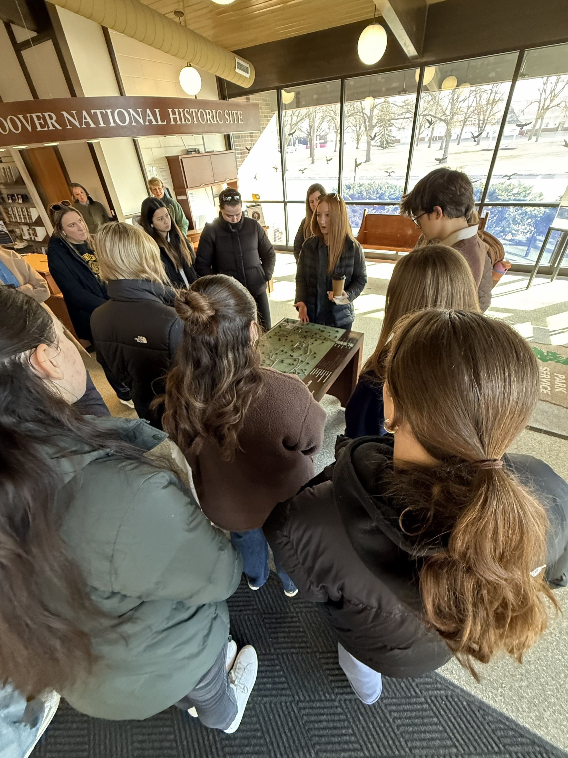A crowd of college students gather around a woman with red hair while she gestures towards a displayed map