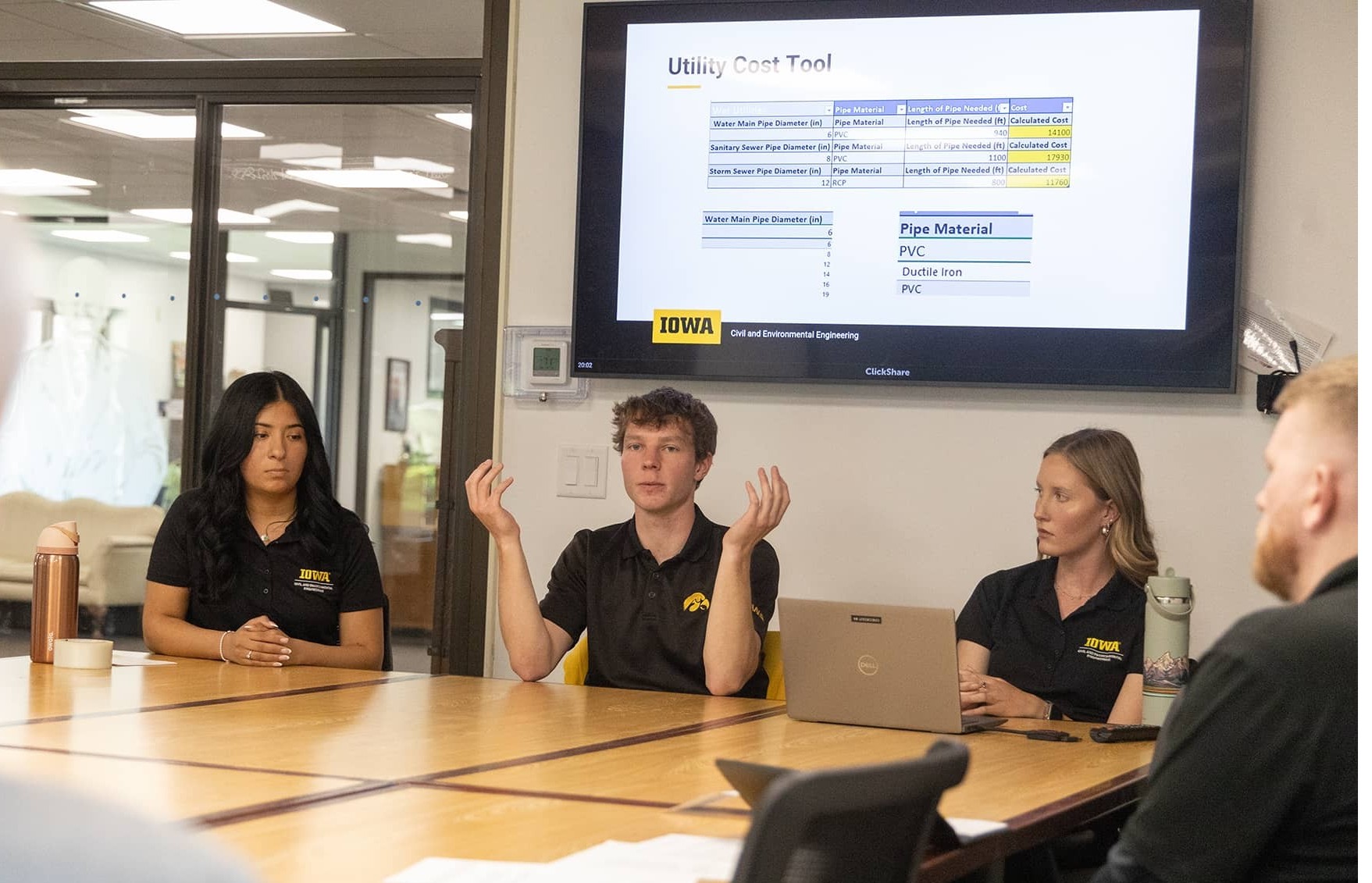 three students sit at a table in front of a slide presentation.