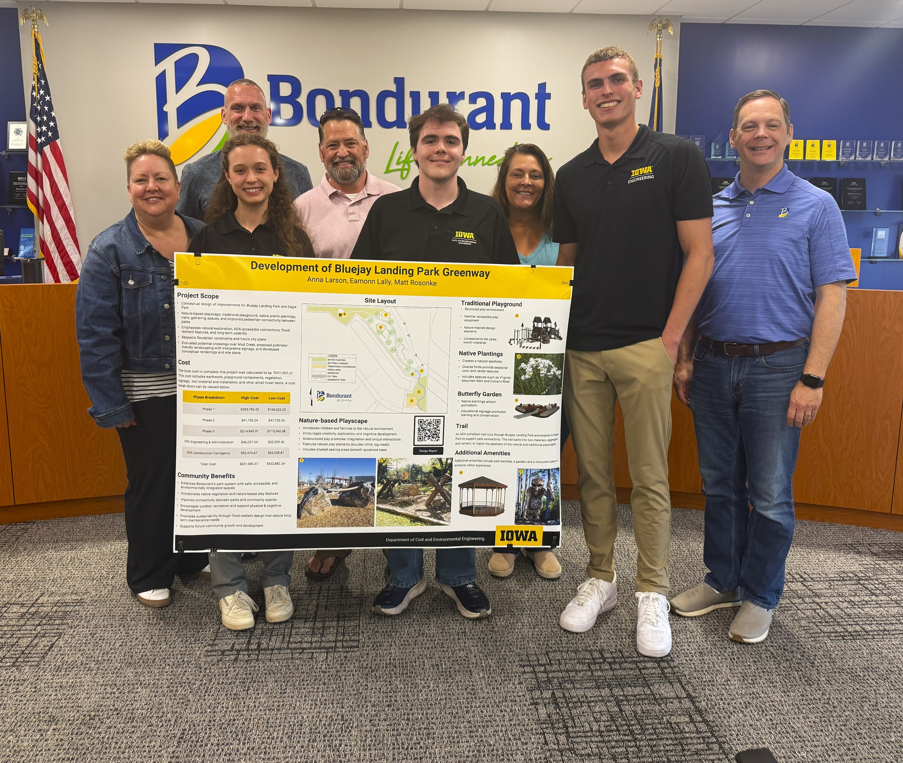 A smiling group of Engineering students, city council members, and the mayor stand in the Bondurant City Council Chambers holding an Engineering poster.