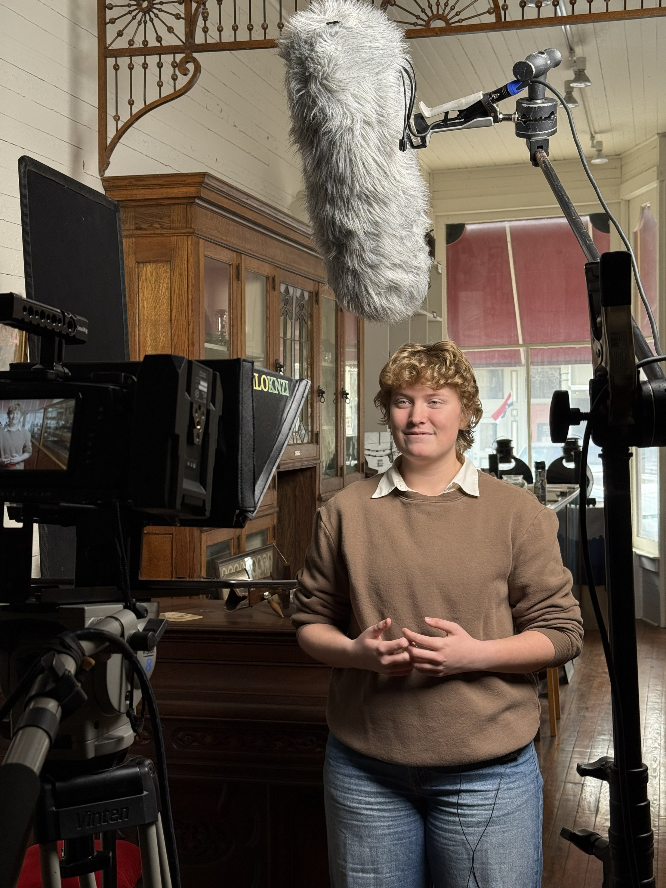 A woman stands speaking with video recording equipment and microphones in front of her.