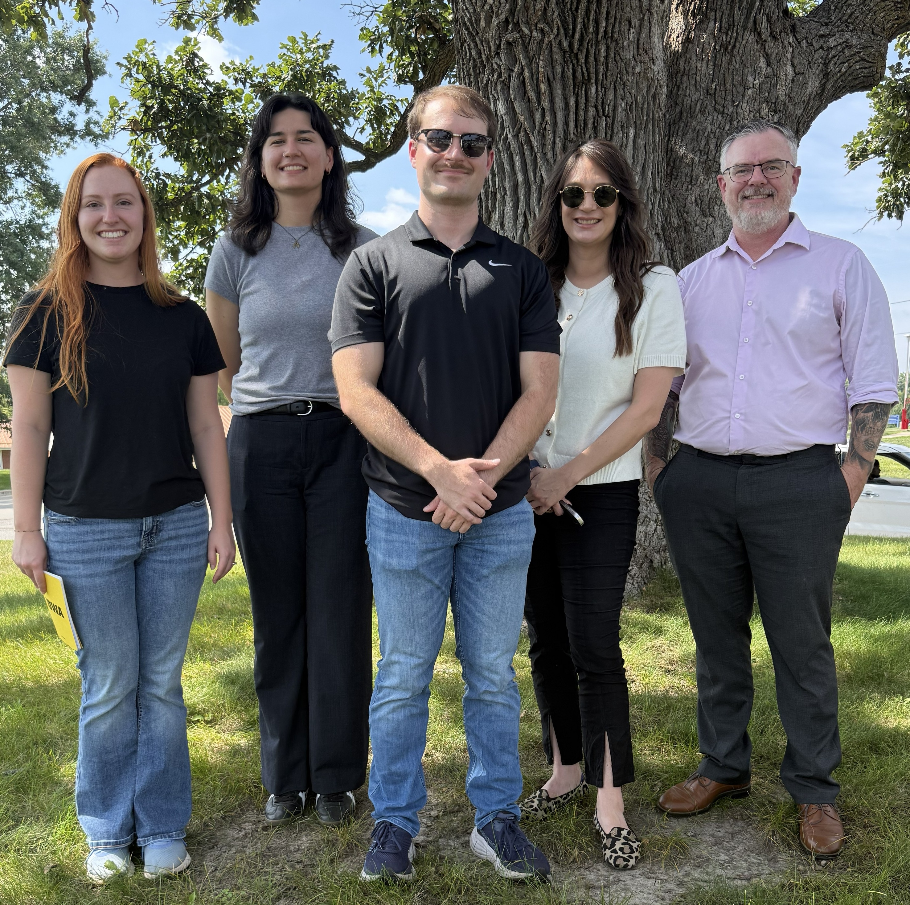 a group of five people stand smiling in front of a tree on a sunny day.