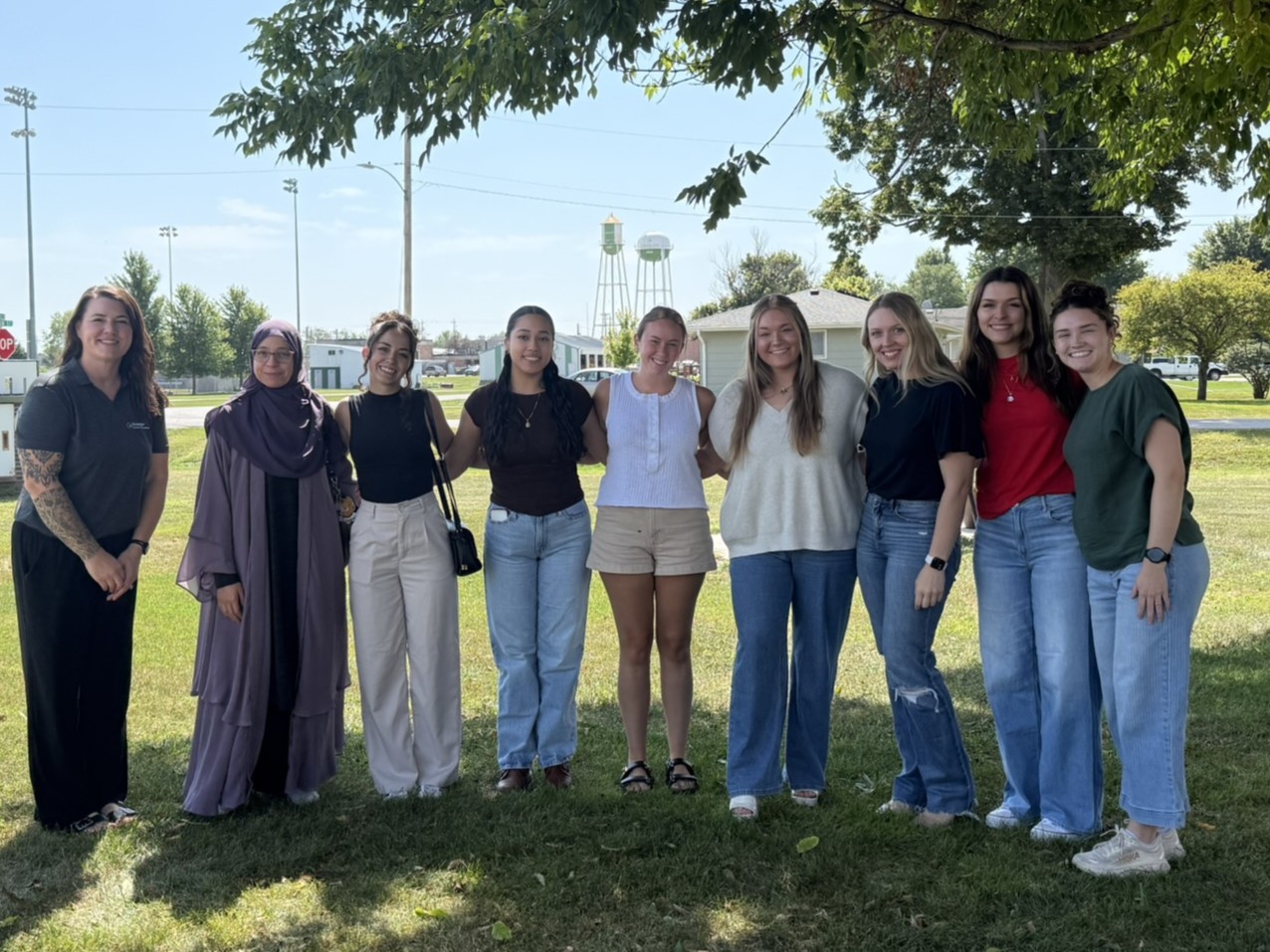 A group of nine people stand outside smiling under a tree with a blue sky.