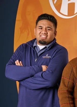 Headshot of middle aged man with brown skin, wearing a purple sweater, arms crossed and smiling