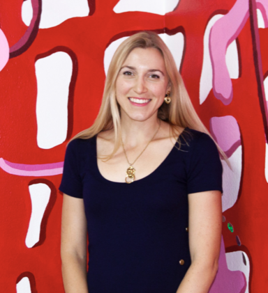 headshot of a young woman with long blonde hair, wearing a black dress and standing in front of a brightly colored wall.