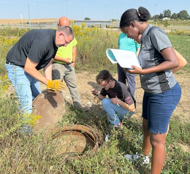 four people cluster around an open manhole cover. One person has a notepad and is watching as a man with gloves gestures towards the hole.