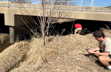 Two people squat on grass in front of a bridge looking down at materials in their hands