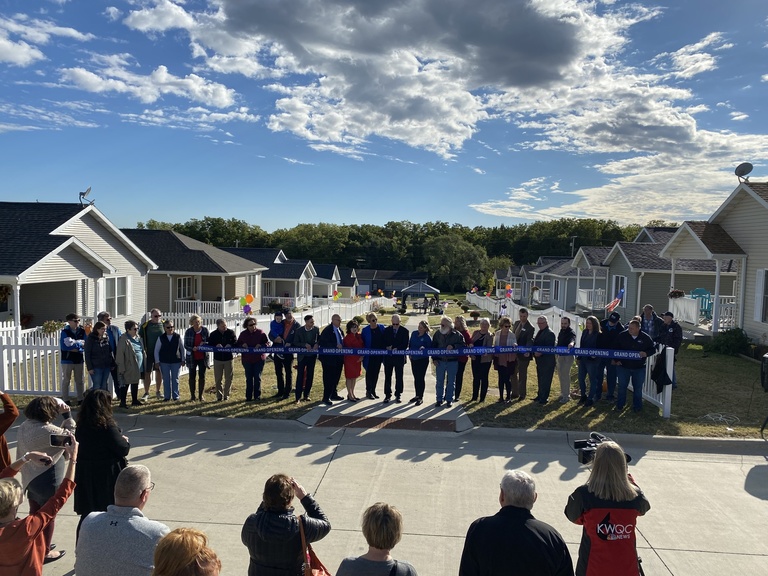A long line of people holding a ribbon and a scissors for a ceremony in front of a group of houses, under a blue sky with clouds.