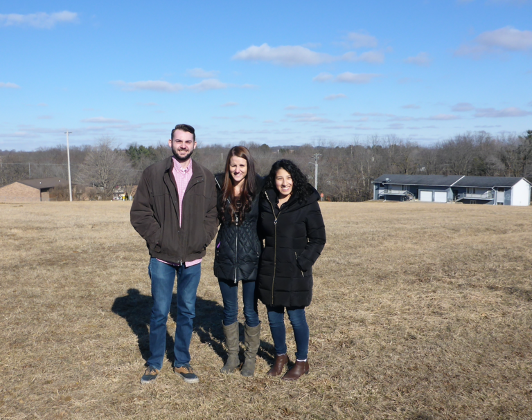 Three young people standing on a bear grassy field and squinting into the sun. 