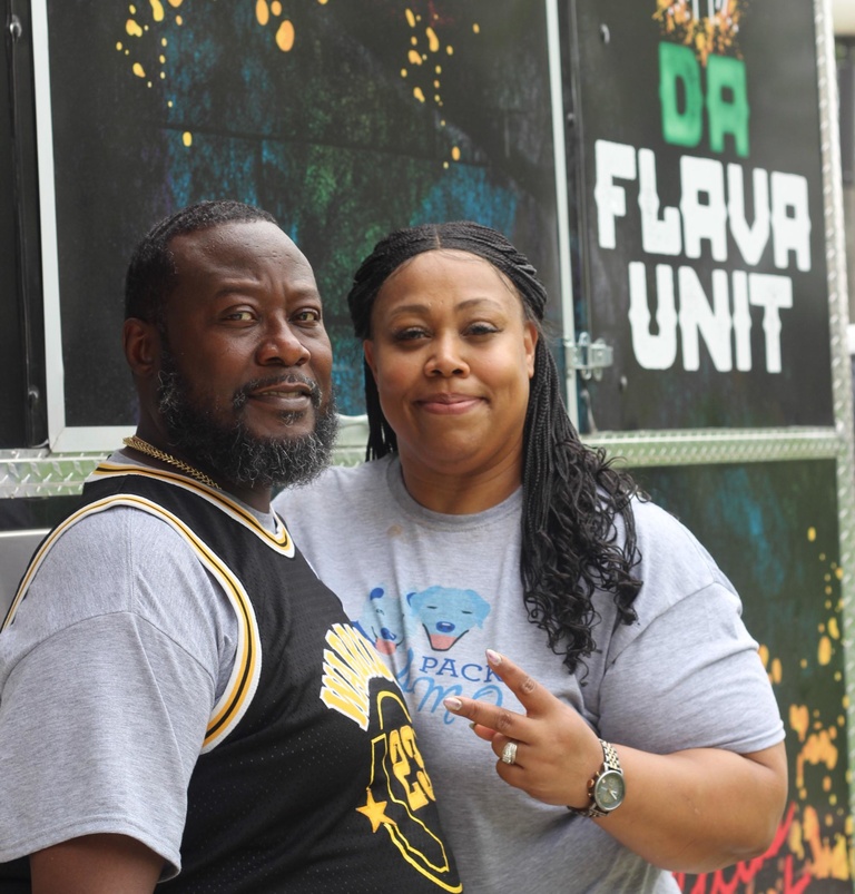 A man and a woman, both of whom are black and wearing casual athletic gear, stand in front of a food truck 
