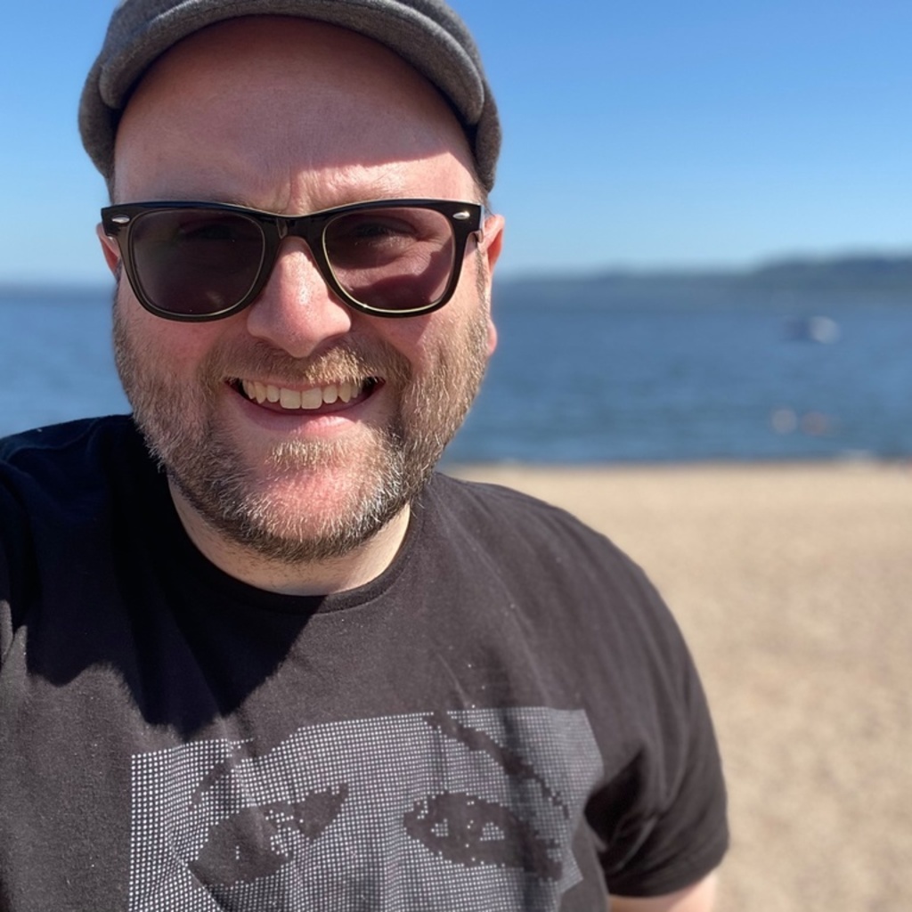 Close up photo of a smiling man wearing a hat and sunglasses on a beach. 