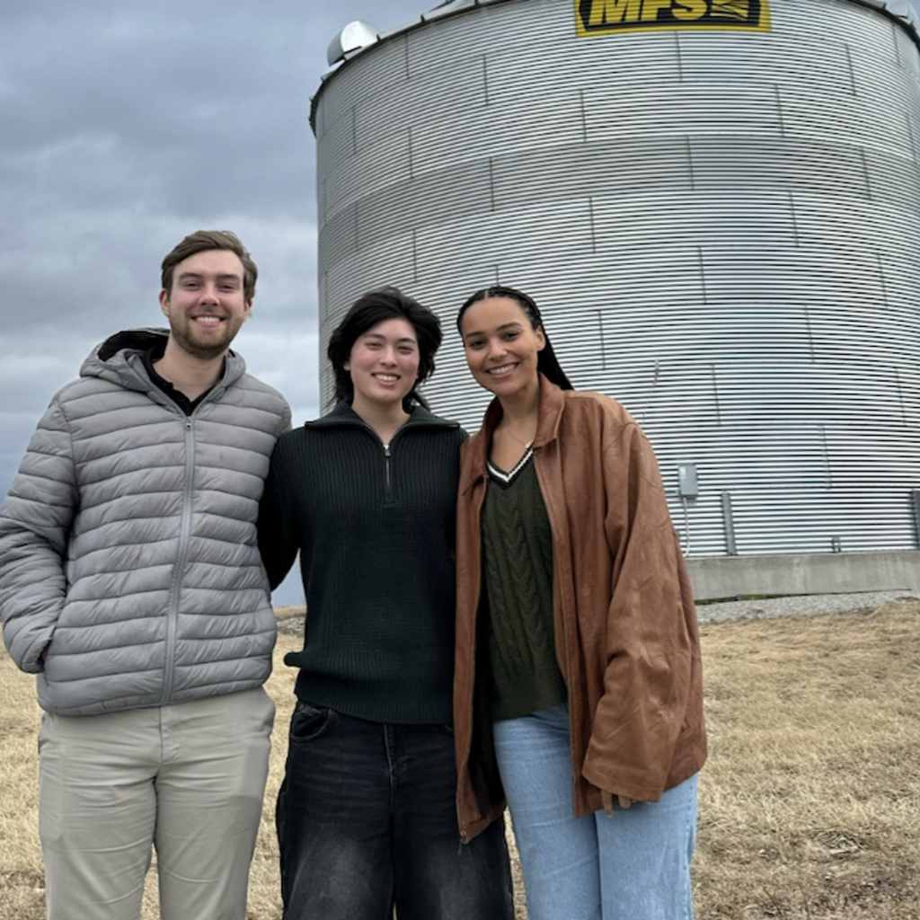 three young people standing in front of a silver metal grain silo on a cloudy day