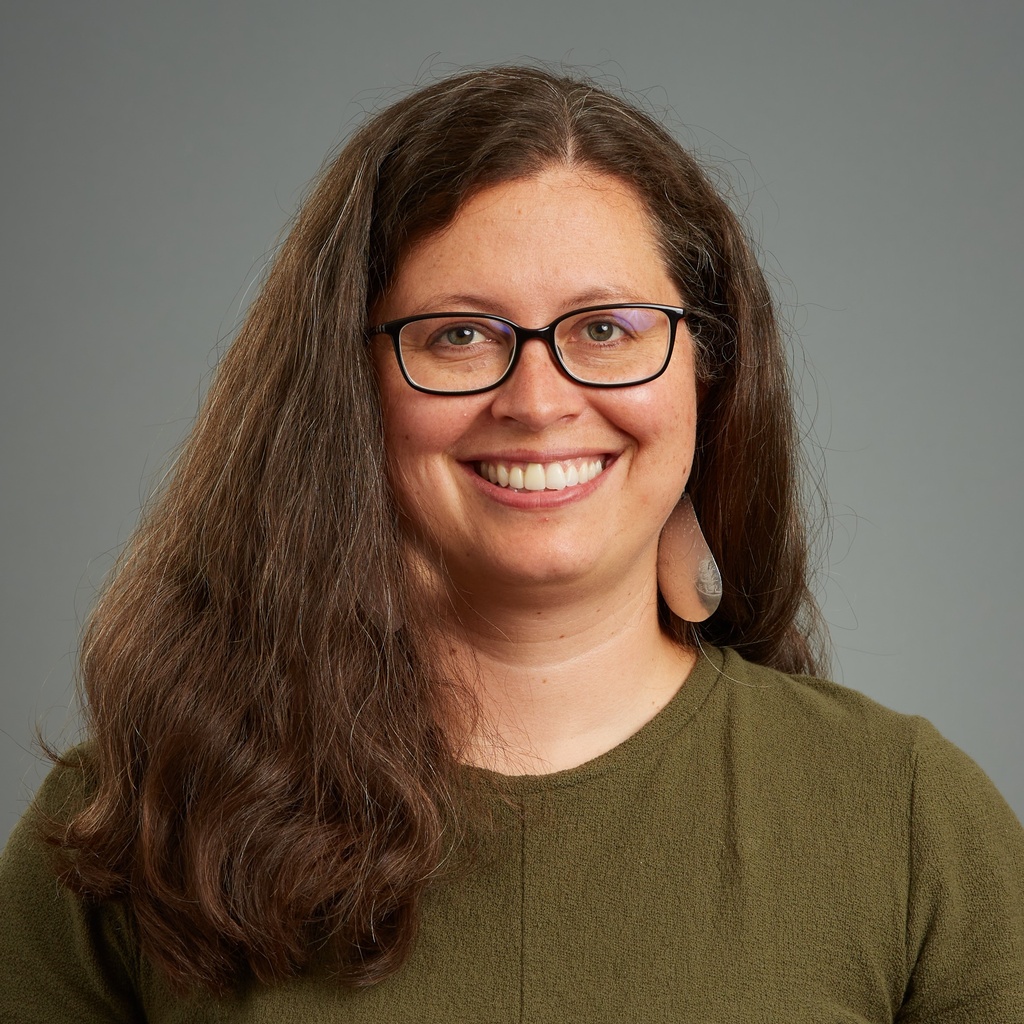 A photo of a white woman with long hair, silver earrings, glasses, and a green shirt.