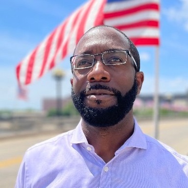Black man in his 40s looking up at the sky with an American flag behind him. He is wearing glasses and a violet dress shirt. 