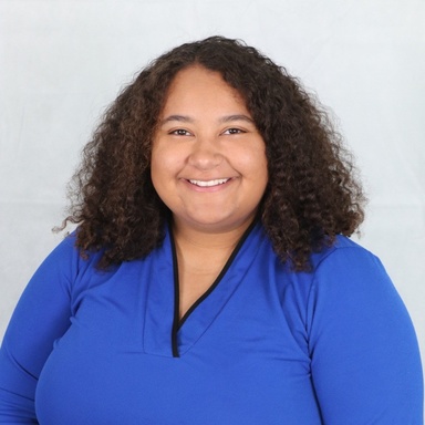 headshot of smiling young woman with long, curly brown hair, brown skin, and a blue shirt.