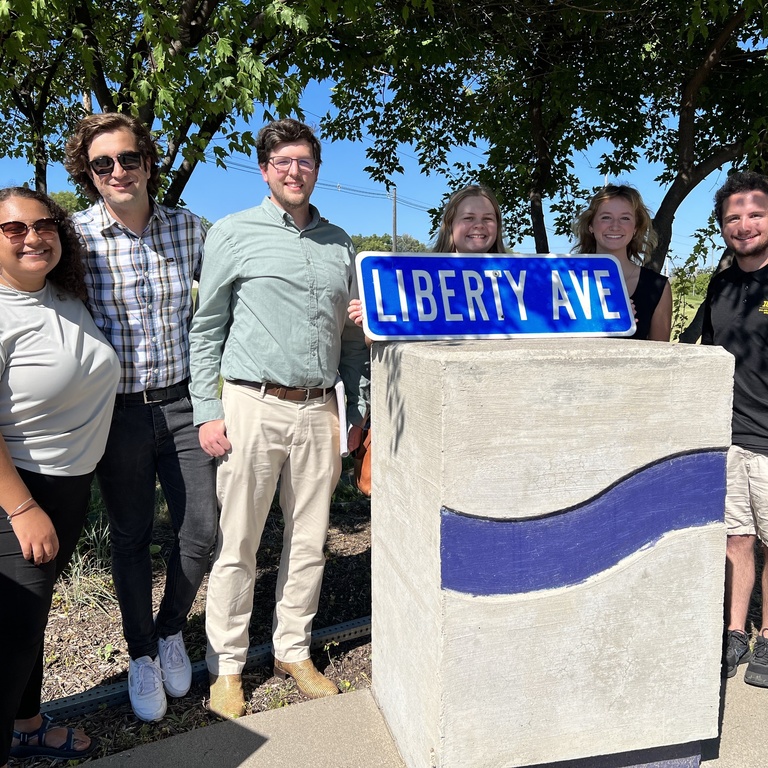 Group of people stands with arms around each other's shoulders in front of a small marker that reads Liberty Ave. 