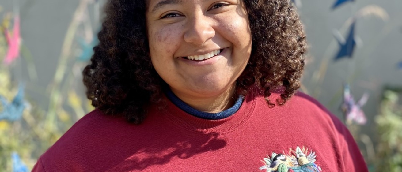 Emani Brinkman, young woman with brown skin, curly shoulder length hair, and wearing a maroon sweater smiles broadly