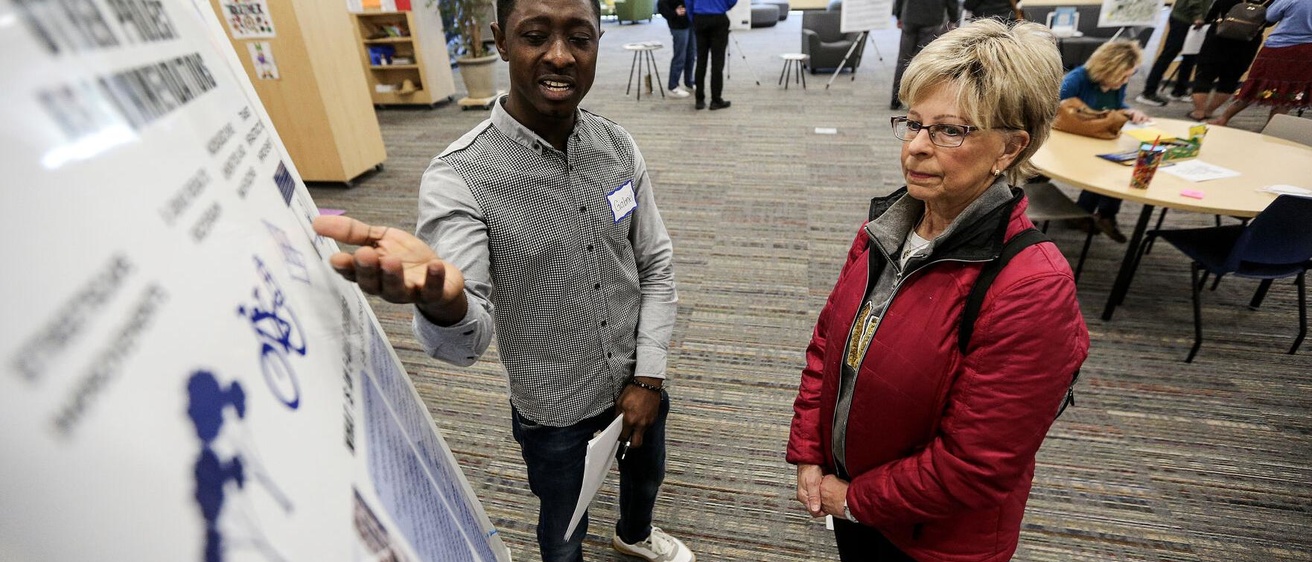 a young man points to a poster and talks to an older woman