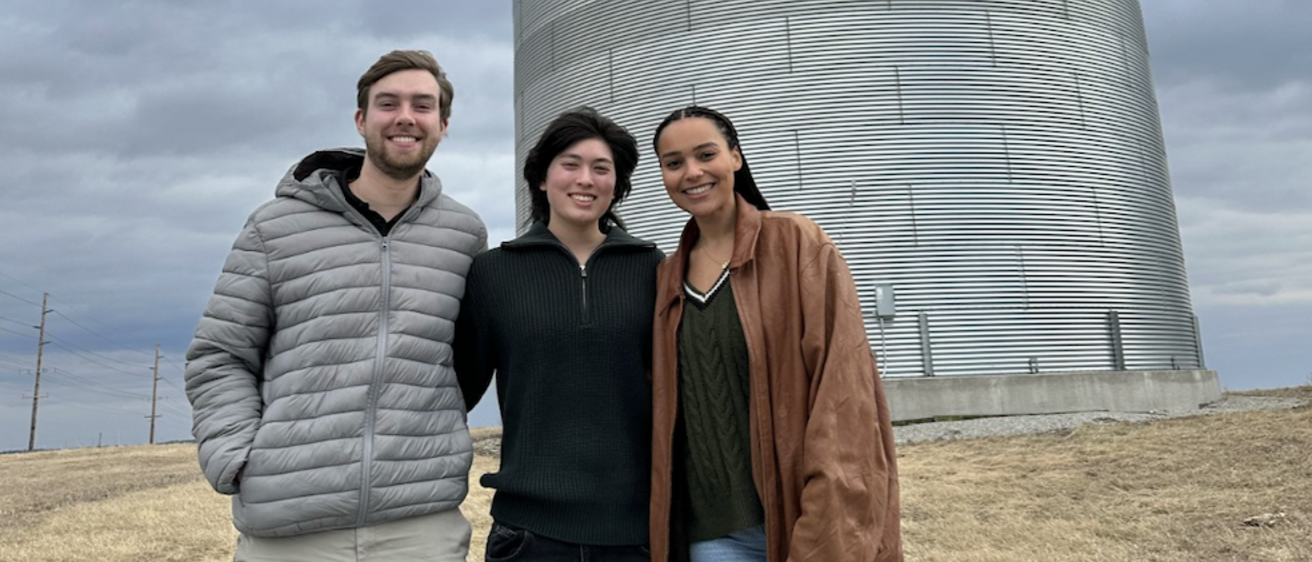 three young people standing in front of a silver metal grain silo on a cloudy day