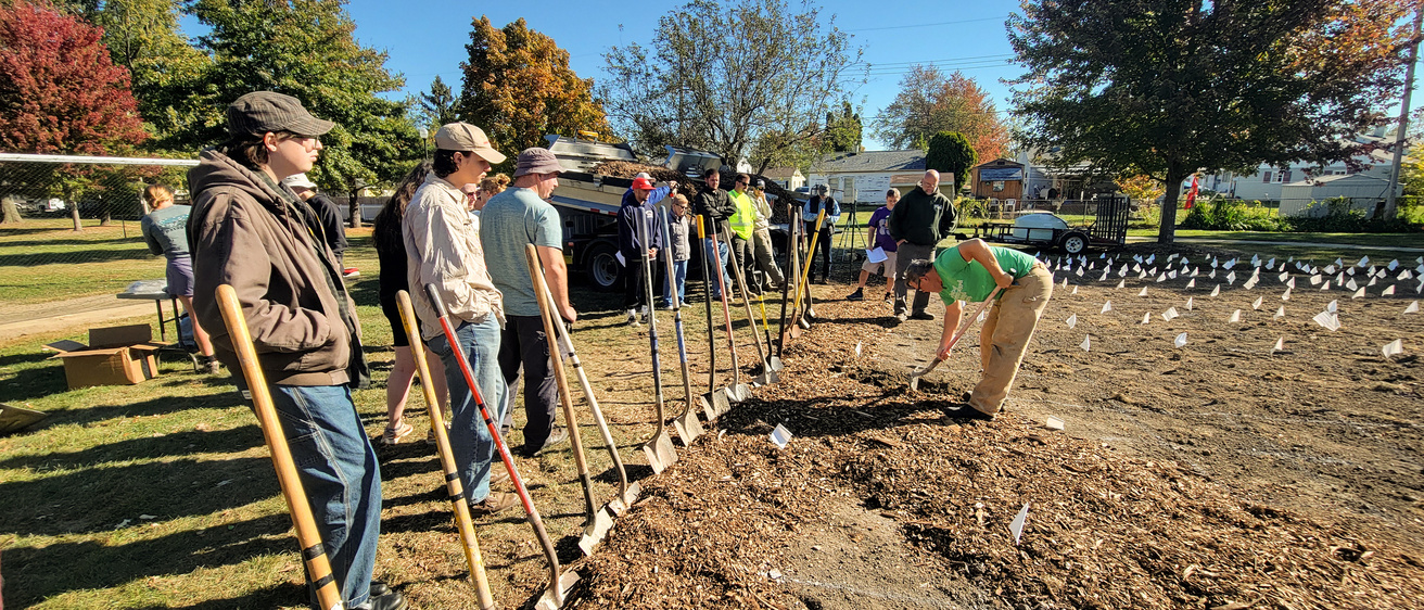 A row of people standing in front of a tilled garden with shovels