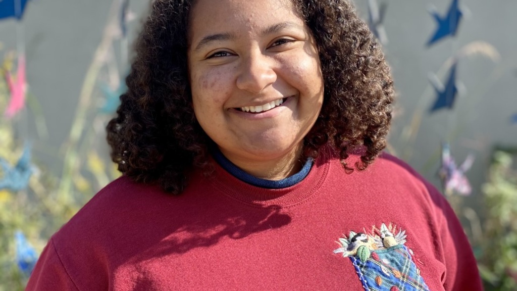 Emani Brinkman, young woman with brown skin, curly shoulder length hair, and wearing a maroon sweater smiles broadly