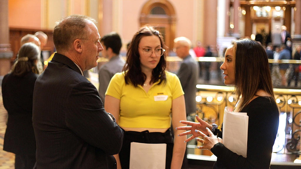 Two young women talk with an older man at a business event with people milling in the background. 