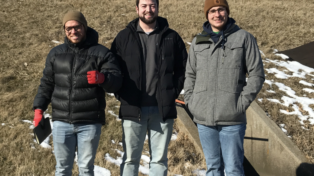 Three young people wearing winter coats and hats stand outside with a small patch of snow and bare ground behind them.