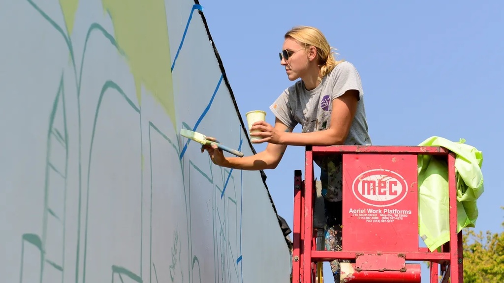 a blonde young woman works on a mural from a crane