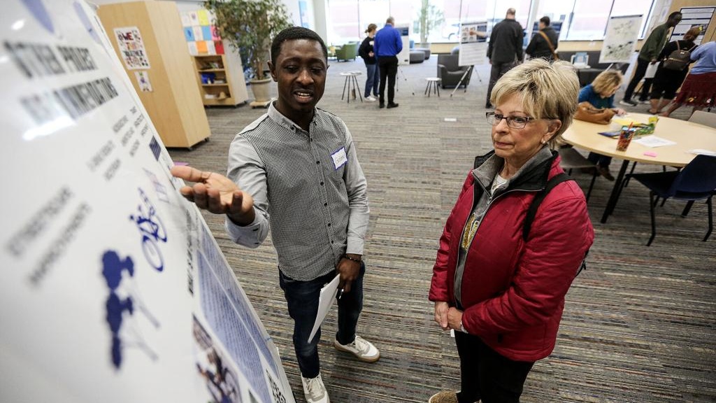 A younger black man and an older white woman talk in front of an informational board to which the man is gesturing.