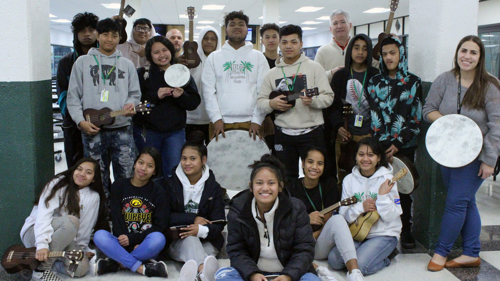A large group of students pose in the hallway of a school with lockers around them. They are smiling and several are holding musical instruments.
