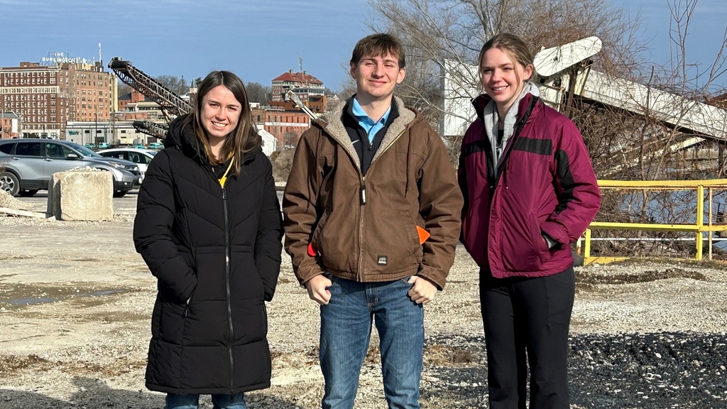 three young people in winter coats standing outside on a sunny day with an industrial site behind them.