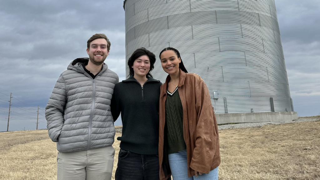 three young people standing in front of a silver metal grain silo on a cloudy day