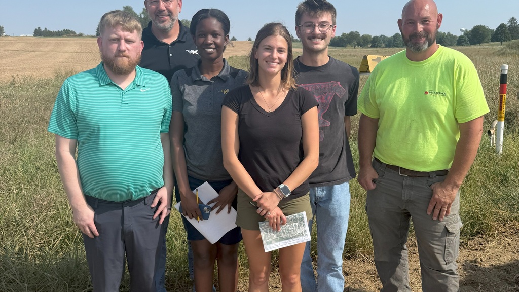 Six people stand on a sunny day in front of a corn field. The three middle people are holding paper maps. The people on each end are wearing West Branch t-shirts.