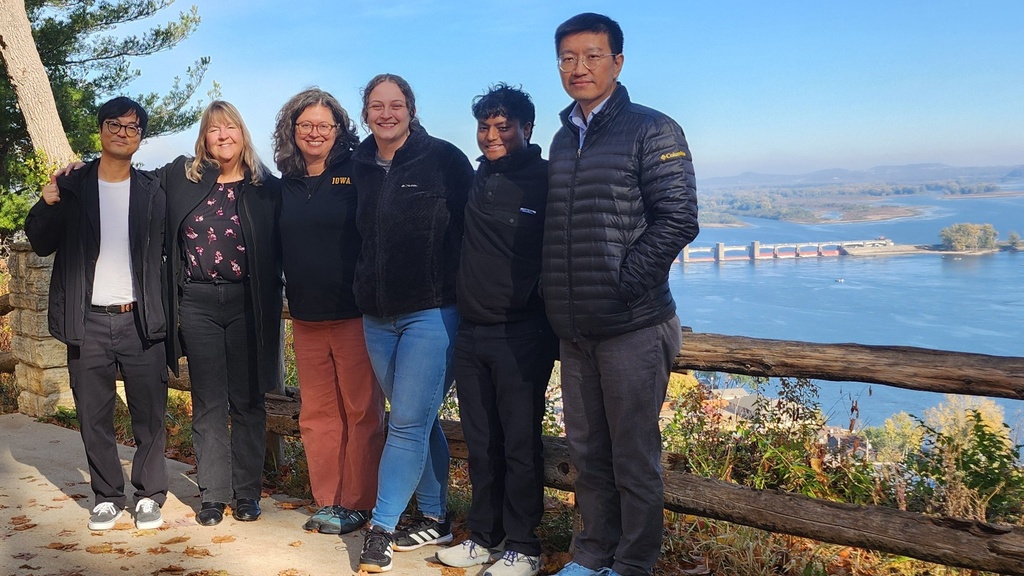 A group of six people stand arm in arm smiling in front of the Mississippi river