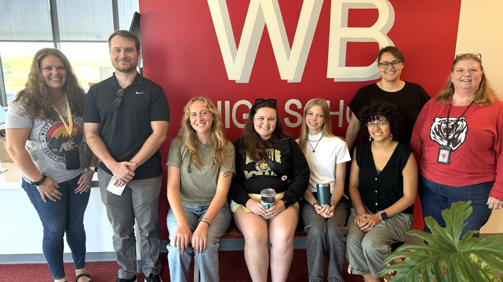 A row of seven people stand and sit smiling in front of a red wall with white letters that says WB