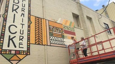 two women on a small crane paint a mural on the side of a building