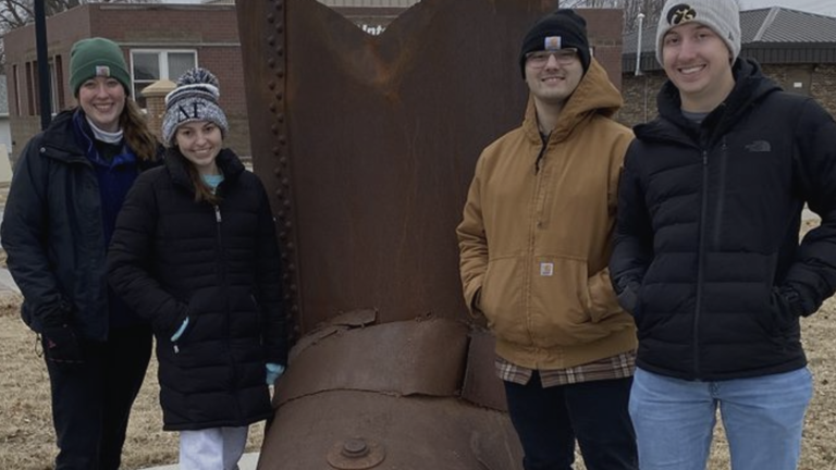Four young adults wearing winter coats and hats stand in front of a bronze sculpture.