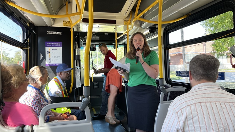 woman in a bright green shirt and blue skirt stands and talks into a microphone on a bus.