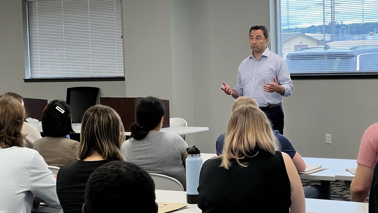 A middle aged white man in a blue shirt talks to a group of people.