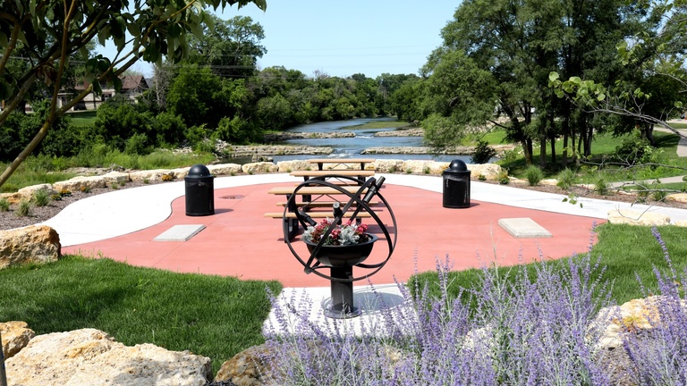 a park on a river with several park benches and a sculpture in the foreground