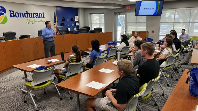 A man in a blue business shirt talks to a group in a lecture hall style room. 