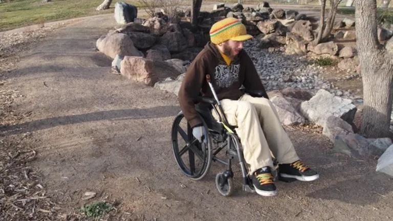 A young man in a yellow hat, dark sweatshirt, and tan pants in a wheelchair on an outdoor dirt path.